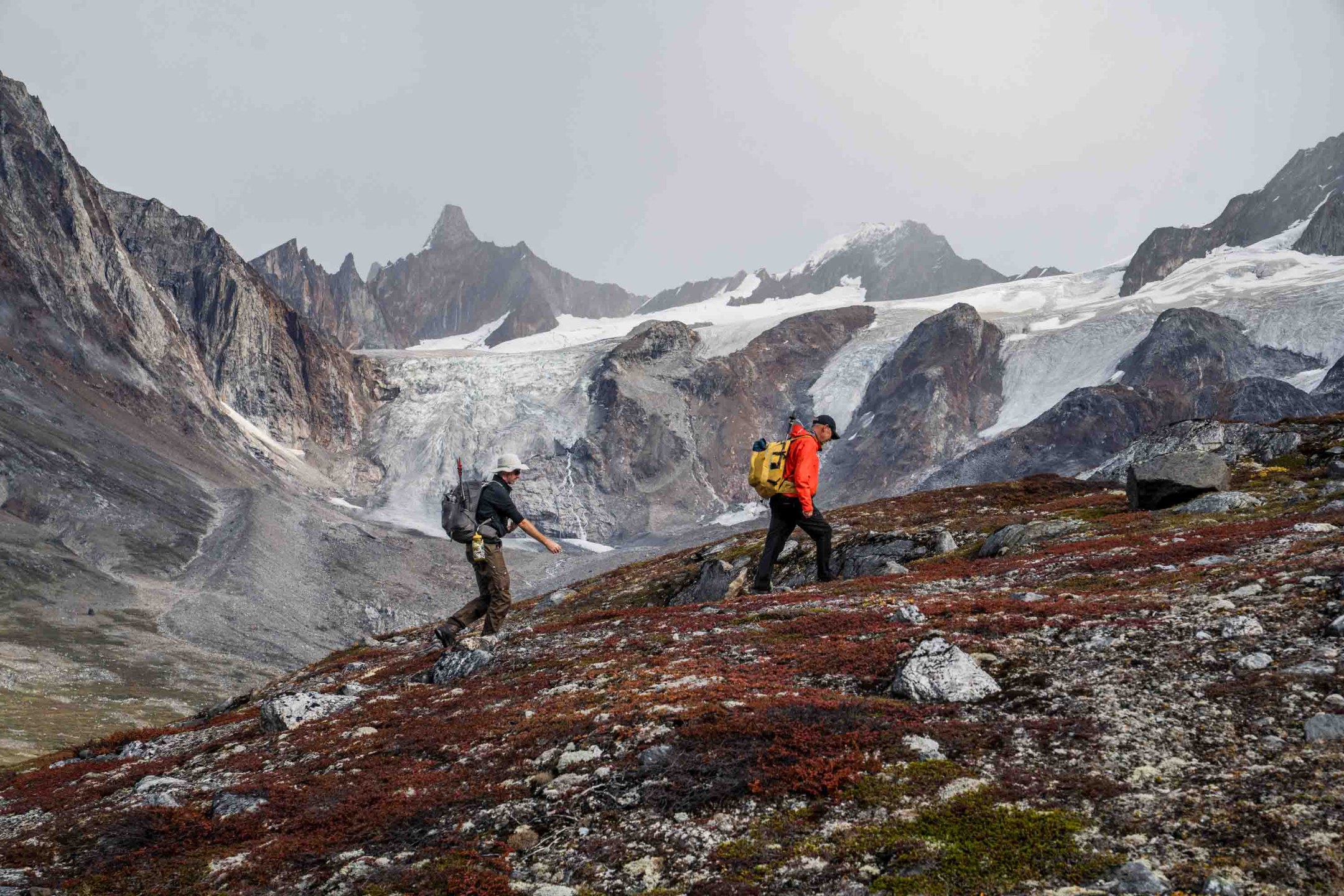 Hikers standing at the edge of a pristine lake in East Greenland, its glassy surface reflecting the dramatic mountains behind them.
