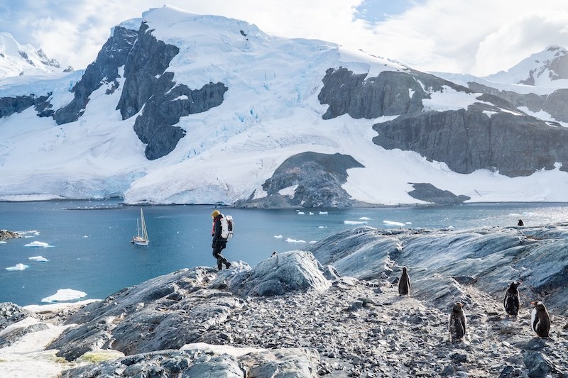 Hiking along rocky Antarctic shoreline with Ocean Tramp expedition vessel anchored offshore and penguins nearby